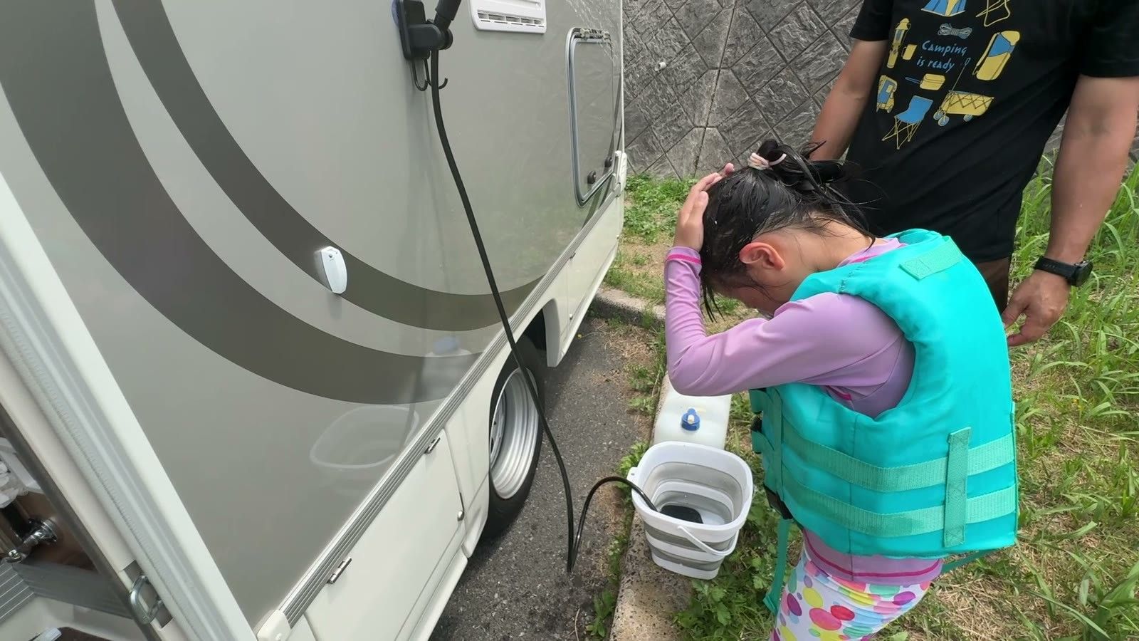 キャンピングカーの横でポータブルシャワーを使って髪を流す娘